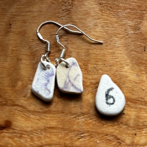 May include: A pair of silver earrings with white ceramic teardrop-shaped charms. One charm has a purple floral design and the other has a white floral design. A separate white ceramic charm with the number 6 is also pictured.