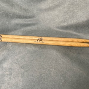 May include: A pair of light brown wooden drumsticks. The drumsticks have the text "UFO Andy Parker" printed on them. The drumsticks are lying on a gray surface.