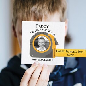 Puede incluir: Una tarjeta blanca con una foto en blanco y negro de un niño sonriendo. La tarjeta dice "Papá, mi amor por ti es inconmensurable." Una cinta métrica amarilla se extiende desde la tarjeta con el texto "¡Feliz Día del Padre! Oliver".