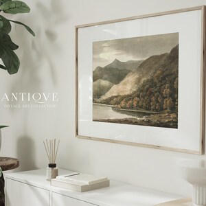 May include: A framed vintage watercolour painting of a lake and mountains. The painting is in a light wood frame and is hanging on a white wall. The text "ANTIQUE VINTAGE ART COLLECTION" is visible below the painting.