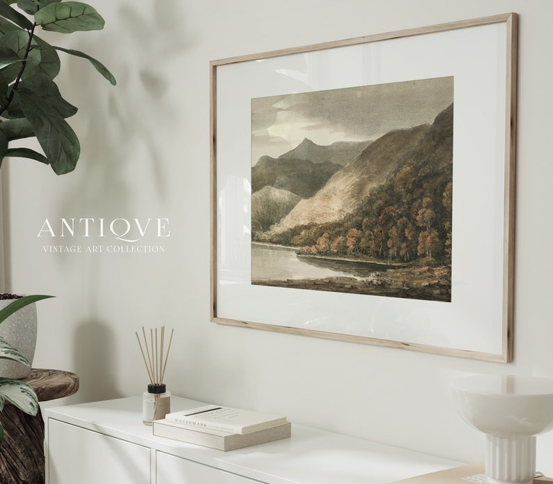 May include: A framed vintage watercolour painting of a lake and mountains. The painting is in a light wood frame and is hanging on a white wall. The text "ANTIQUE VINTAGE ART COLLECTION" is visible below the painting.