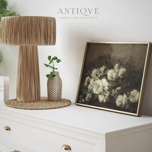 May include: A white dresser with two drawers and gold knobs. On top of the dresser is a brown woven lamp with a fringe shade, a small white vase with a geometric pattern, and a framed print of white roses in a dark background.