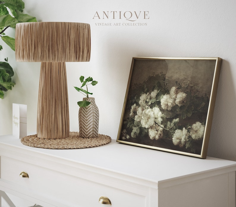 May include: A white dresser with two drawers and gold knobs. On top of the dresser is a brown woven lamp with a fringe shade, a small white vase with a geometric pattern, and a framed print of white roses in a dark background.
