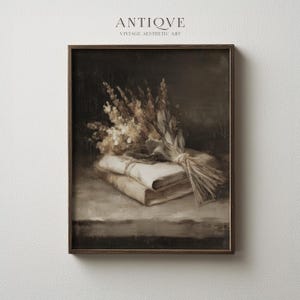 Puede incluir: Una pintura de naturaleza muerta en tonos sepia que presenta un ramo de flores secas atadas con hilo, descansando sobre dos libros antiguos. La obra de arte está enmarcada en un marco de madera oscura, con las palabras "ANTIQVE" y "VINTAGE AESTHETIC ART" en la parte superior.
