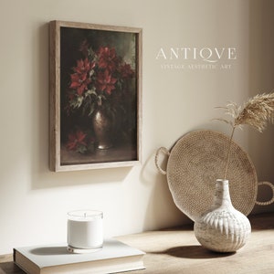 May include: A framed painting of a bouquet of red poinsettias in a silver vase. The painting is hanging on a light beige wall. The wall is decorated with a woven tray, a white vase with a dried grass stem, and a white candle in a glass jar.