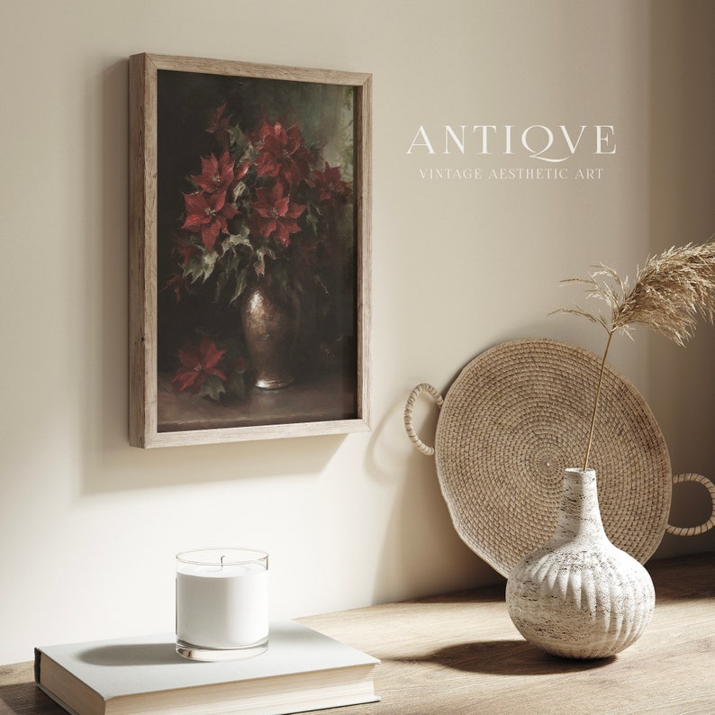 May include: A framed painting of a bouquet of red poinsettias in a silver vase. The painting is hanging on a light beige wall. The wall is decorated with a woven tray, a white vase with a dried grass stem, and a white candle in a glass jar.