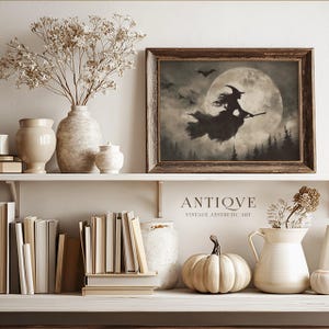May include: A framed vintage print depicts a witch flying on a broom in front of a full moon, with bats in the sky. The print is displayed on a shelf with books, vases, and a pumpkin. The text "ANTIQUE VINTAGE AESTHETIC ART" is below the print.