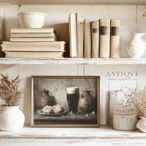 May include: A framed print of a still life painting depicting a glass of dark beer, two ceramic jugs, and two pastries on a wooden table. The painting is in a gold frame and is displayed on a white shelf with other decorative items.