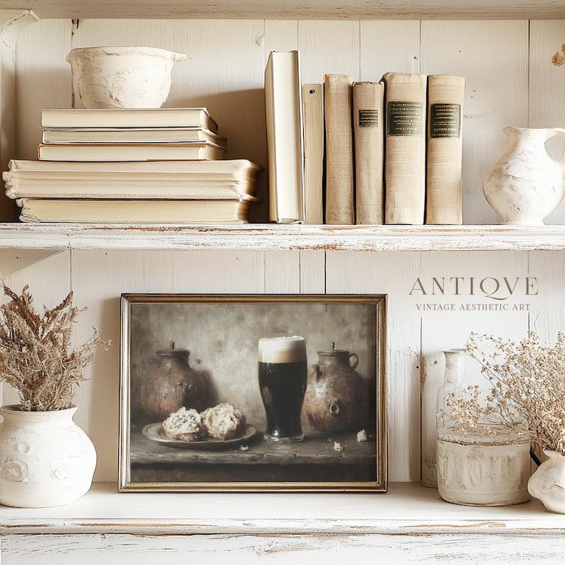 May include: A framed print of a still life painting depicting a glass of dark beer, two ceramic jugs, and two pastries on a wooden table. The painting is in a gold frame and is displayed on a white shelf with other decorative items.