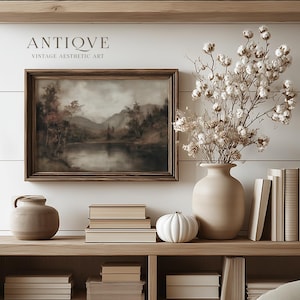 May include: A framed landscape painting with a wooden frame, depicting a lake scene with trees and mountains. The text "ANTIQVE VINTAGE AESTHETIC ART" is above the painting. A beige vase holds dried flowers, and a white pumpkin sits on a wooden shelf with books.