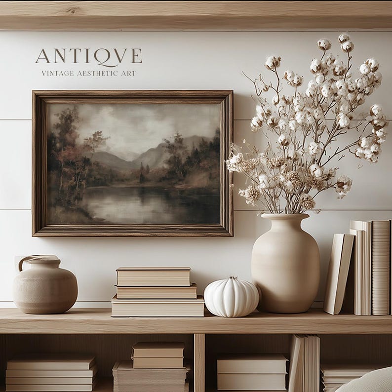 May include: A framed landscape painting with a wooden frame, depicting a lake scene with trees and mountains. The text "ANTIQVE VINTAGE AESTHETIC ART" is above the painting. A beige vase holds dried flowers, and a white pumpkin sits on a wooden shelf with books.