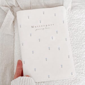 May include: A white rectangular notebook cover featuring a repeating pattern of small light blue tulips. The text "Mutterpass" is printed in black, followed by the cursive script "growing love" centred on the front cover.