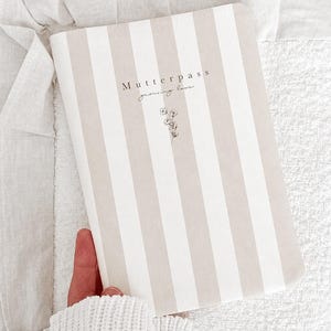 May include: A rectangular book cover featuring a vertical striped pattern in beige and white. The text "Mutterpass" and "growing love" is printed in black, accompanied by a small, delicate line drawing of a flower centred below the text.