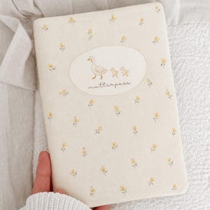 May include: A rectangular fabric cover in cream with a small yellow floral pattern. Centered is an oval patch featuring an illustration of a mother duck and two ducklings with the text "mutterpass" written in a cursive script below the birds.