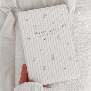 May include: A rectangular notebook with a light grey and white vertical striped cover featuring small brown floral sprigs. The text "Mutterpass" and "growing love" is printed in the centre in a dark font. The cover has rounded corners.
