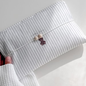 Könnte beinhalten: Eine rechteckige Stofftasche mit vertikalem Streifenmuster in Weiß und Grau. Die Tasche wird mit einer dünnen weißen elastischen Kordel, zwei hellen Holzperlen und einem kleinen braunen Teddybären-Anhänger verschlossen. Der Text "little love" ist aufgedruckt.
