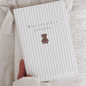 Könnte beinhalten: Ein rechteckiges Notizbuch mit weißem Einband und dünnen, vertikalen hellblauen Streifen. Der Text "Mutterpass" und "growing love" ist in Dunkelbraun gedruckt, ergänzt durch eine kleine Illustration eines braunen Teddybären in der Mitte.