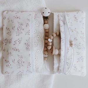 Könnte beinhalten: Eine weiße Reißverschlusstasche mit Blumenmuster und weißem Spitzenbesatz. Die Tasche ist geöffnet und enthält einen Holz-Beißring, eine Holzperlenkette und einen Schnuller.