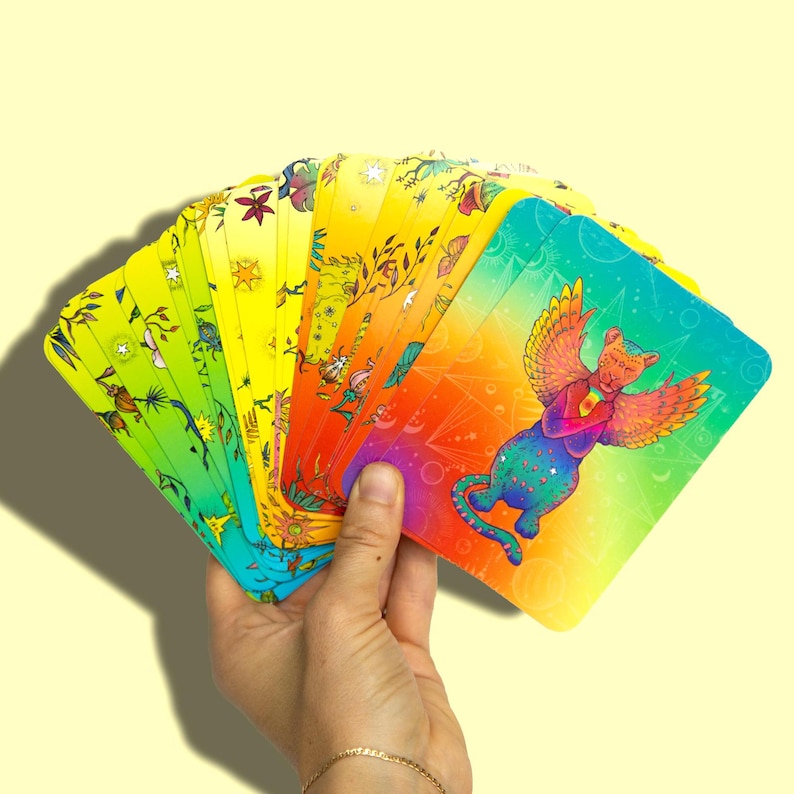 May include: A deck of colourful oracle cards featuring a rainbow cat with wings. The cards have a yellow background with a variety of illustrations and symbols.