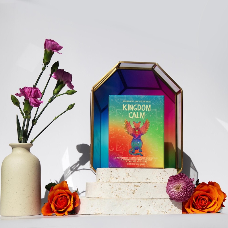 May include: A deck of cards with a rainbow background and a colourful creature on the front. The text on the front of the cards reads "Kingdom Calm". The cards are displayed on a white tiered stand with flowers on either side.