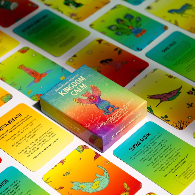 May include: A box of Kingdom Calm cards with a colourful rainbow design and a mythical creature on the front. The box is surrounded by cards with colourful illustrations and text.