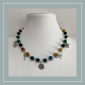 Peut inclure: Un collier de perles compos&eacute; de pierres rondes multicolores dans des tons de vert, bleu et marron. Le collier est agr&eacute;ment&eacute; de breloques en m&eacute;tal argent&eacute;, notamment une spirale, une cl&eacute;, un bouton, une libellule et une forme d'&eacute;toile.