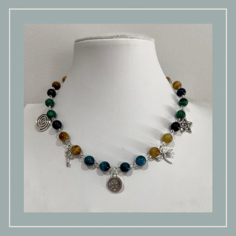 Peut inclure: Un collier de perles compos&eacute; de pierres rondes multicolores dans des tons de vert, bleu et marron. Le collier est agr&eacute;ment&eacute; de breloques en m&eacute;tal argent&eacute;, notamment une spirale, une cl&eacute;, un bouton, une libellule et une forme d'&eacute;toile.