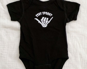 May include: A black short-sleeved baby bodysuit featuring a white graphic print of a skeletal hand making a shaka sign gesture. Above the skeletal hand, the text STAY SPOOKY is printed in a bold, white, sans-serif font on the chest area.