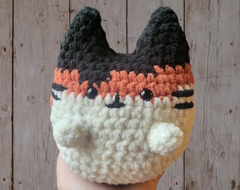 May include: A crocheted stuffed animal shaped like a cat with black, orange, and white fur. The cat has black eyes and a white nose.