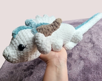 May include: White crocheted dragon plush toy with blue and brown accents. The dragon has a long tail and a small, cute face.