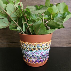 May include: A terracotta flower pot with a colorful fabric rope wrapped around it. The pot is filled with green leafy plants.