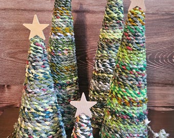May include: Five cone-shaped decorative trees wrapped in multicolored fabric strips in shades of green, yellow, and blue. Each tree is topped with a light-colored wooden star. The trees vary in height and are arranged in a cluster.