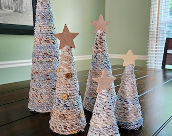 May include: A set of five cone-shaped decorative trees wrapped in multicolored fabric strips. Each tree is topped with a wooden star. One tree features several brown buttons attached to its surface. The trees vary in height and are arranged together.
