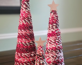 May include: Three cone-shaped decorative trees of varying heights, each wrapped in a textured fabric cord featuring a mix of red, white, and black colors. Each tree is topped with a light-colored wooden star ornament fixed to the peak.