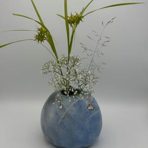 Könnte beinhalten: Eine runde, hellblaue Keramikvase mit gesprenkelter Oberfläche und feinen weißen Streifen. Die Vase enthält ein Blumenarrangement aus kleinen weißen Schleierkrautblüten, langen grünen Grashalmen und stacheligen grünen Samenkapseln.