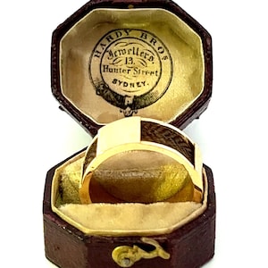 Op de afbeelding: Een gouden ring met een houten inlay in een rode achthoekige doos. De doos heeft een label met de tekst "Hardy Bros. Jewellers 13 Hunter Street SYDNEY."