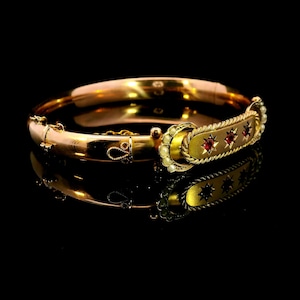 May include: A gold-coloured hinged bangle bracelet with a rectangular centrepiece. The centrepiece features three red gemstones and is bordered by small pearls. The bracelet is displayed on a reflective black surface.