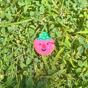 May include: A pink and green painted strawberry with a smiling face, lying on a bed of green grass.