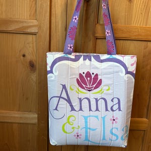 May include: A quilted tote bag with a light purple and white floral design. The bag features the names "Anna & Elsa" in purple and blue lettering, with a pink flower graphic. The handles are purple with pink flower accents.