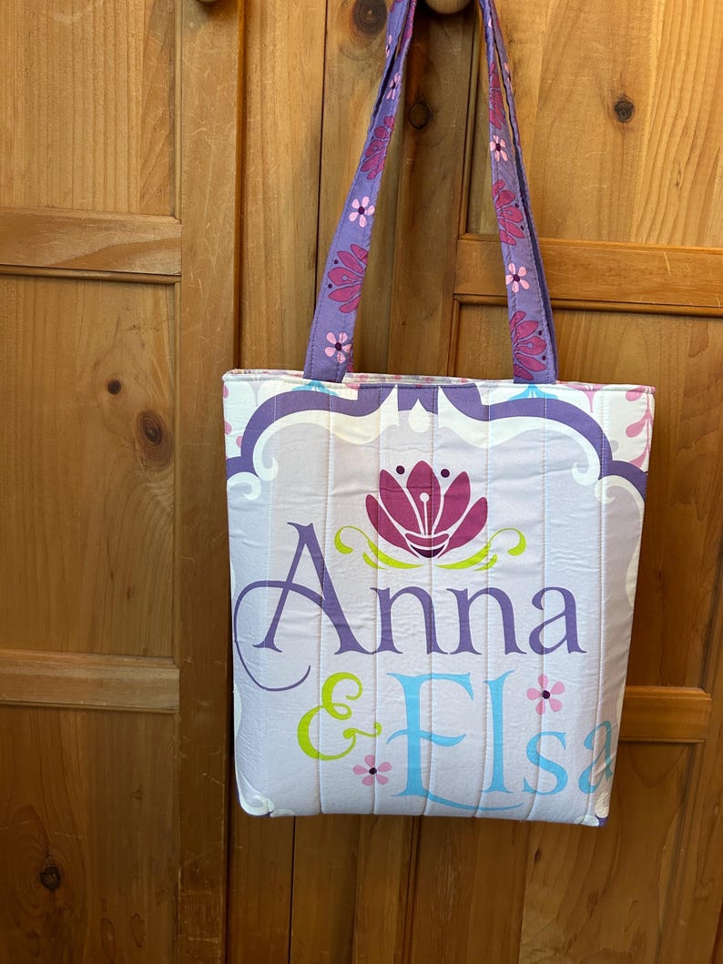 May include: A quilted tote bag with a light purple and white floral design. The bag features the names "Anna & Elsa" in purple and blue lettering, with a pink flower graphic. The handles are purple with pink flower accents.