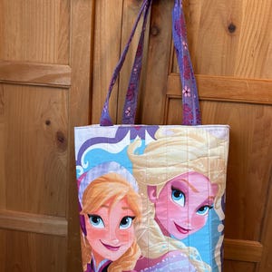 May include: Quilted tote bag featuring a colorful illustration of two animated characters with blonde hair and blue eyes. The bag has purple straps with a floral pattern and a quilted design. The background is a light blue and white color.