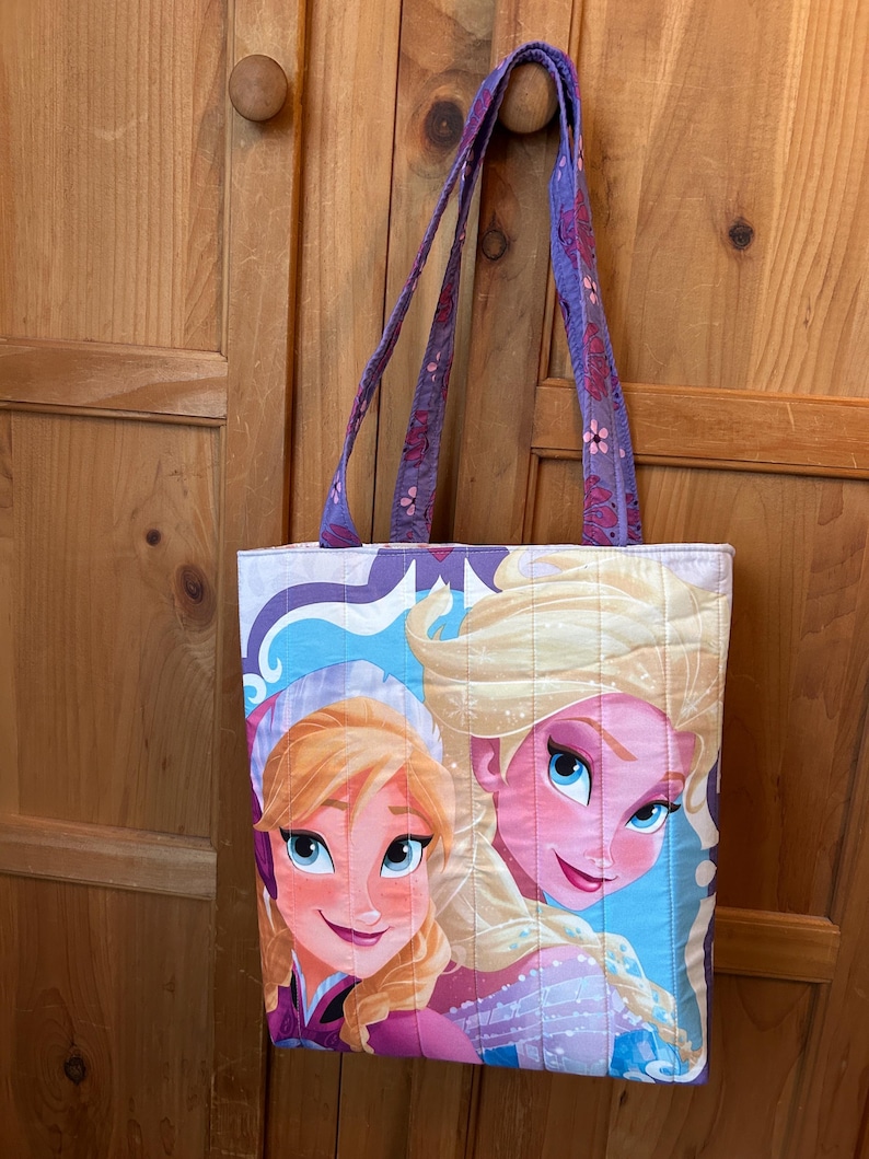 May include: Quilted tote bag featuring a colorful illustration of two animated characters with blonde hair and blue eyes. The bag has purple straps with a floral pattern and a quilted design. The background is a light blue and white color.