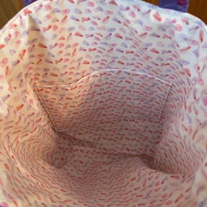May include: A purple tote bag with a white interior patterned with red and purple brushstrokes. The bag has a small pocket inside. The handles are purple with a floral design.