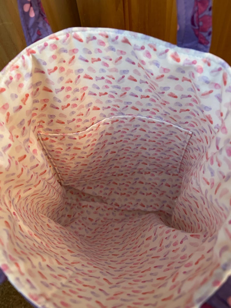 May include: A purple tote bag with a white interior patterned with red and purple brushstrokes. The bag has a small pocket inside. The handles are purple with a floral design.
