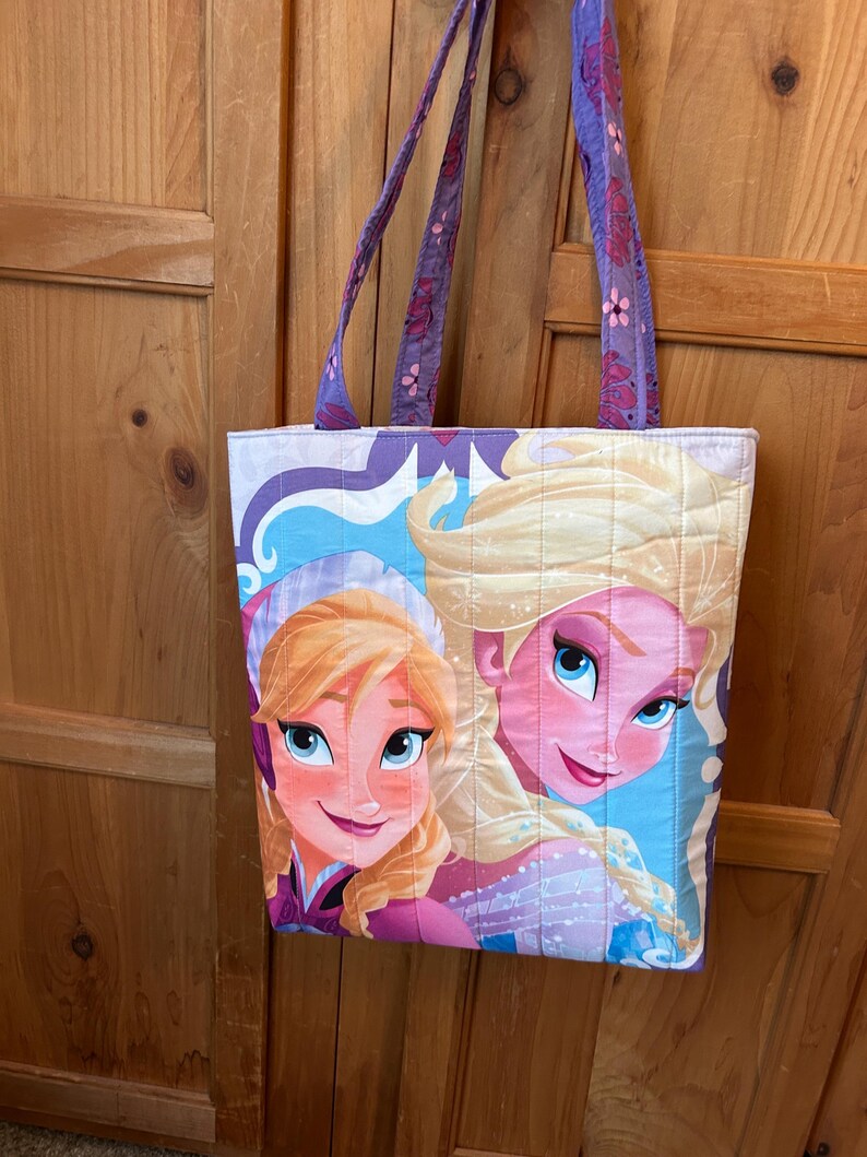 May include: Quilted tote bag featuring a colorful illustration of two animated characters with blonde hair and blue eyes. The bag has purple straps with a floral pattern and a quilted design. The background is a light blue and white pattern.