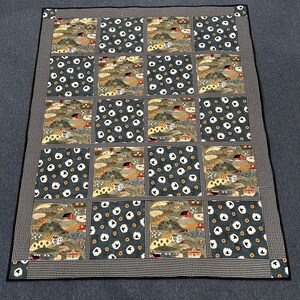 May include: A patchwork quilt with a repeating pattern of landscape scenes and white circles on a dark blue background. The quilt is bordered with a black and white checkered fabric.