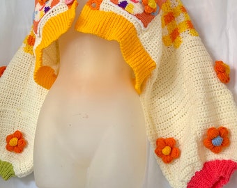 Puede incluir: Un bolero de crochet color crema con detalles en naranja, rosa y amarillo. El bolero presenta un diseño floral con flores naranjas y un ribete amarillo brillante. El texto "Usahana Crochet Pattern" es visible en la parte inferior.