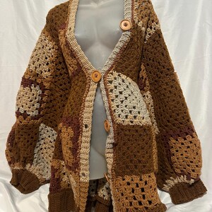 以下が含まれることがあります： Vネックとボタン留めで、茶色、ベージュ、クリーム色のクロシェ編みのカーディガン。カーディガンはパッチワークの正方形でできており、ゆったりとしたオーバーサイズのフィット感です。