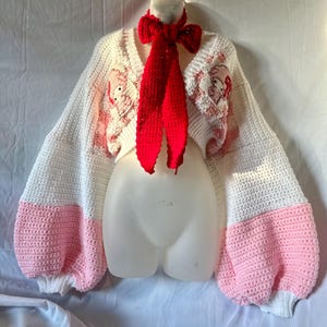 May include: A white and pink crochet cardigan with long, puffy sleeves. The sleeves are white with pink cuffs. The cardigan has a V-neck and features a red crochet scarf. The cardigan has a floral design on the chest and sleeves.