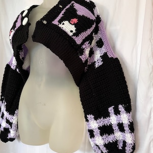 Op de afbeelding: Een zwart en paarse gebreide cardigan met een wit en zwart geruit patroon op de mouwen. De cardigan heeft een getailleerde lengte en een V-hals. De cardigan is op een wit mannequin. "Kuromi Crochet Pattern" staat in zwarte tekst op een witte achtergrond onder het mannequin.